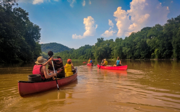 Three canoes are paddled away from the camera by young people wearing life jacket. The river is muddy and lined by thick trees. 